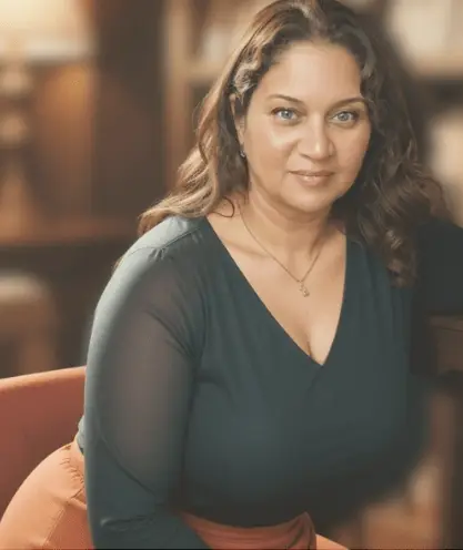 Smiling woman with wavy brown hair wearing a dark green blouse, seated in a warmly lit library setting with a softly blurred background.