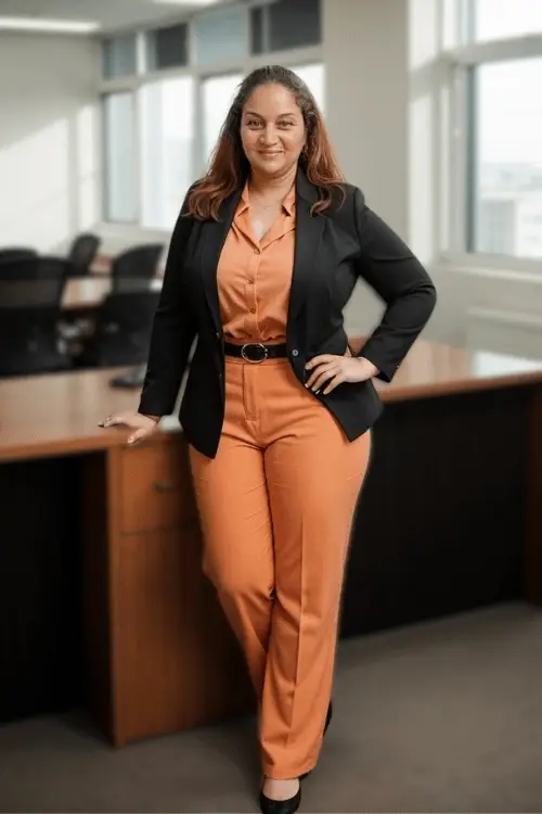 Professional woman in a rust-colored suit and black blazer standing confidently in a modern office. The background is softly blurred, keeping the focus on her poised stance and warm expression.