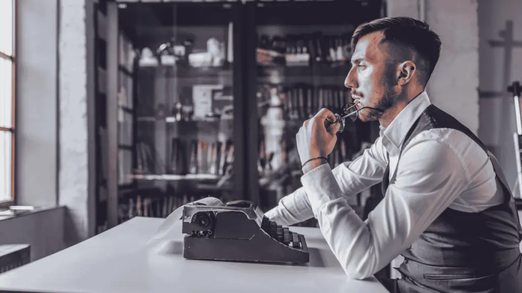 A pensive male writer, sitting at a desk, holding his glasses in one hand.