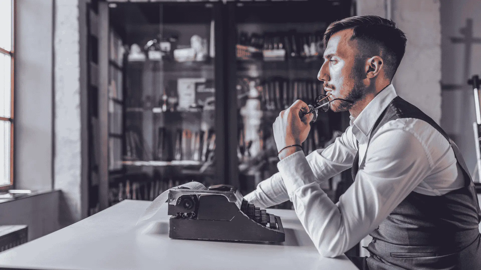 A pensive male writer, sitting at a desk, holding his glasses in one hand.